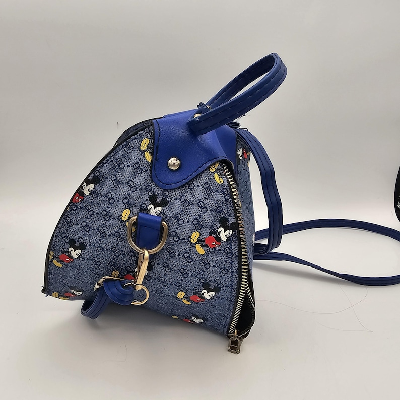 May include: A blue triangular handbag featuring a repeating pattern of Mickey Mouse figures and a small logo print. The bag includes a solid blue top handle, a silver-toned metal zip closure, and a detachable blue shoulder strap with gold-toned hardware.