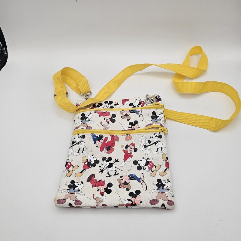 May include: A white crossbody bag featuring a repeating pattern of Mickey Mouse in various poses and outfits. The bag has two zippered compartments with yellow zips and a matching yellow adjustable shoulder strap made of woven fabric.