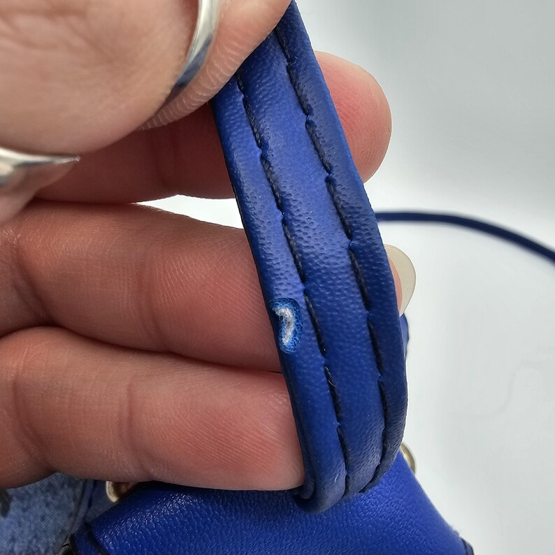 May include: A bright blue handbag strap featuring a textured surface and dark blue stitching along the edges. The strap displays a small area of visible wear where the blue material has been scuffed, revealing a lighter colour underneath the surface.