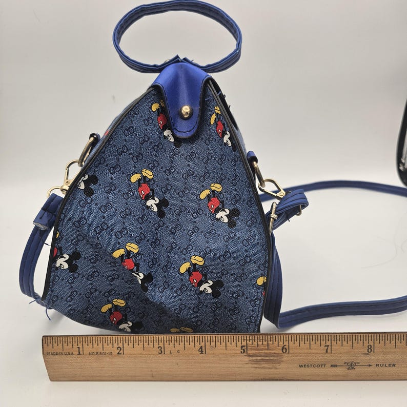 May include: A triangular blue handbag featuring a repeating pattern of Mickey Mouse and the letters GG. The bag has a top handle, gold-tone hardware, and a detachable shoulder strap. A wooden ruler placed at the base shows the bag is 20.3 cm wide.