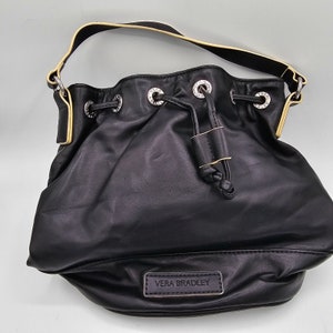 May include: A black bucket bag with a drawstring closure, silver-tone metal grommets, and a single shoulder strap featuring light yellow piping. The front displays a rectangular black patch with the embossed text VERA BRADLEY in silver.