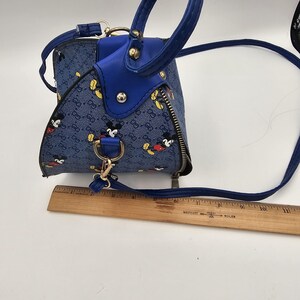 May include: A blue handbag with a repeating Mickey Mouse pattern and a monogram print, featuring solid blue trim, gold-tone hardware, and a zipper closure. A wooden ruler placed underneath shows the bag is approximately 23 cm in length.