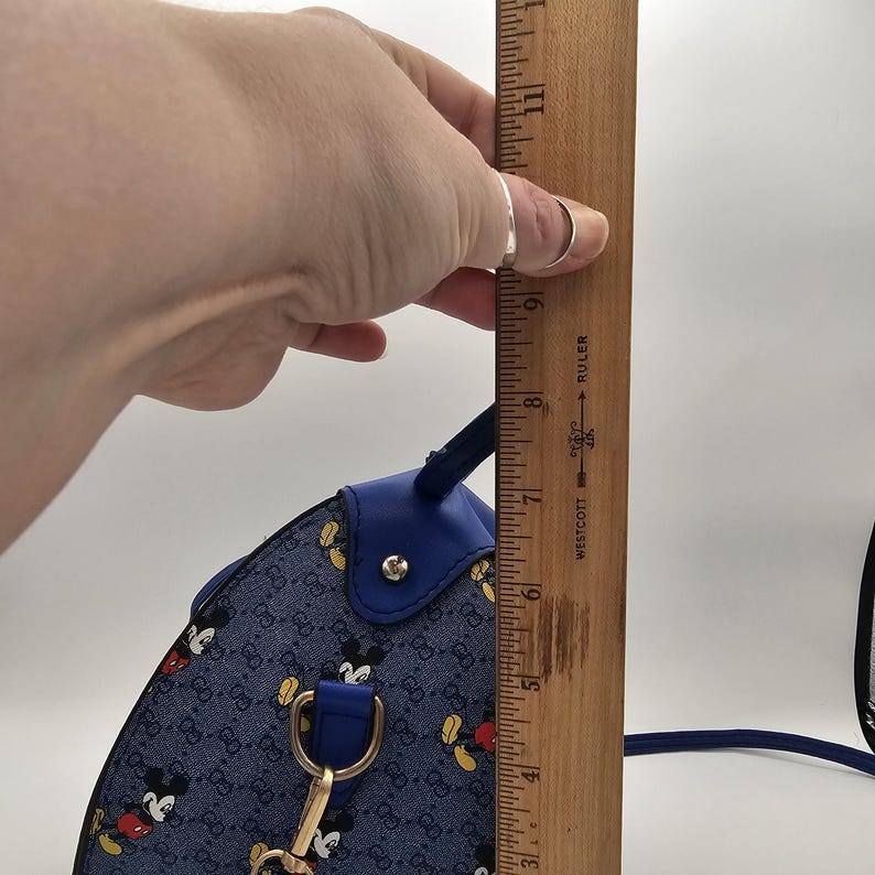 May include: A blue denim handbag featuring a repeating Mickey Mouse pattern and a gold-tone metal hardware attachment. A wooden Westcott ruler is positioned vertically against the side of the bag, showing a height measurement of approximately 15 cm.