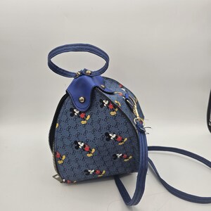 May include: A blue handbag with a repeating pattern of Mickey Mouse figures and a small interlocking logo design. The bag features a rounded top handle, a side zipper closure, and a thin blue shoulder strap attached with gold-toned metal hardware.