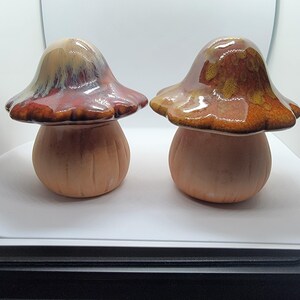 May include: Two ceramic mushroom sculptures with smooth, matte, light tan stems and glossy, rounded caps. The left cap features a blend of deep red, brown, and cream glaze, while the right cap displays a mottled amber and brown glaze finish.