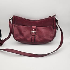 May include: A deep burgundy leather shoulder bag featuring a curved top zipper closure, a front exterior pocket with a vertical strap, and a silver-toned metal buckle accent. The bag includes an adjustable shoulder strap attached with silver hardware.