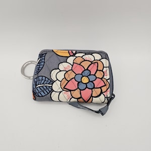 May include: A rectangular quilted coin purse in a grey fabric featuring a large floral pattern in shades of pink, peach, cream, and blue. The accessory includes a silver metal key ring attached to the side and a grey fabric zipper pull tab.