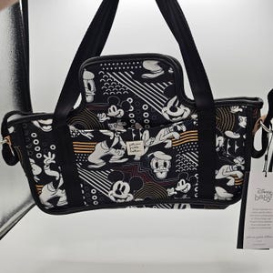 May include: A black and white Petunia Pickle Bottom bag featuring a repeating Disney character print of Mickey Mouse, Donald Duck, and Goofy. The bag has black fabric handles, a central pocket with a brand label, and a white Disney Baby hang tag.