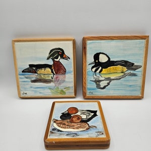 May include: Three square wooden-framed tiles featuring hand-painted duck illustrations. Two tiles depict ducks swimming in blue water, while the third shows two ducks resting. Each tile is signed with the name Jim in the corner of the artwork.