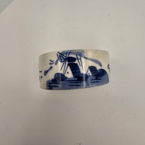May include: A ceramic napkin ring with a blue and white design. The design features a landscape with trees and a windmill. The napkin ring is cylindrical and has a slightly curved shape.