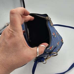 May include: A blue crossbody bag featuring a repeating pattern of Mickey Mouse characters. The bag has a top zip closure with a metallic pull, a dark interior lining, and a thin blue shoulder strap attached with brass-toned metal hardware.