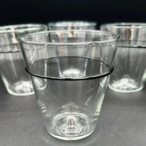 May include: A set of four clear glass tumblers, each featuring a distinct black horizontal line encircling the upper portion of the vessel. The glasses have a slightly tapered shape with a thick, reinforced base that includes a small, raised center point.