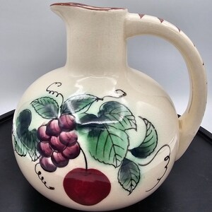 May include: A cream-colored ceramic pitcher with a round body and a handle. The pitcher is decorated with a hand-painted design of grapes and a cherry on a vine.