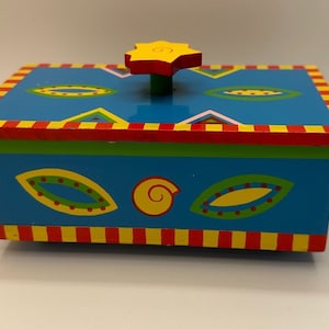 May include: A rectangular wooden box with a bright blue base and lid. The lid features a yellow star-shaped knob with a spiral center. The box is decorated with yellow and green leaf-like shapes, red spirals, and red and yellow striped borders.