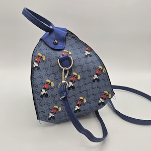 May include: A blue backpack featuring a repeating pattern of cartoon mouse characters and a small interlocking letter motif. The bag has a top handle, blue trim, and a detachable shoulder strap attached with a silver-toned metal lobster clasp.