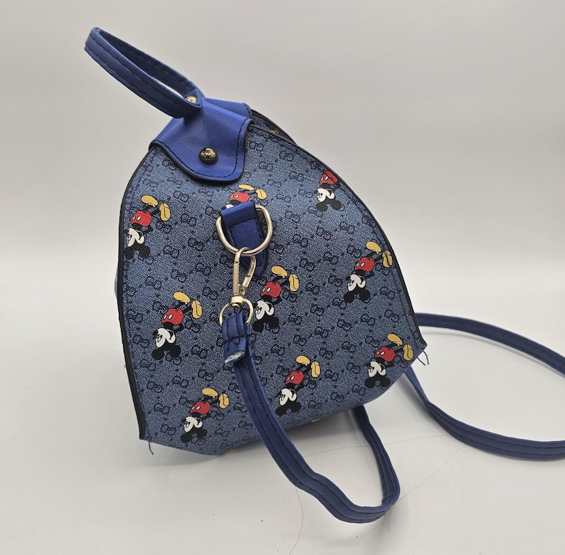 May include: A blue backpack featuring a repeating pattern of cartoon mouse characters and a small interlocking letter motif. The bag has a top handle, blue trim, and a detachable shoulder strap attached with a silver-toned metal lobster clasp.