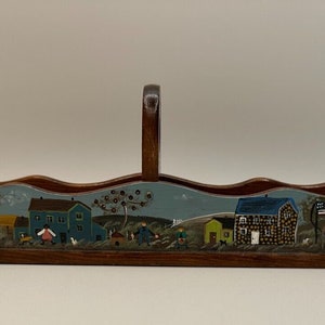May include: A wooden serving tray with a painted scene of houses and people. The tray has a handle and is painted in shades of blue, green, brown, and yellow.