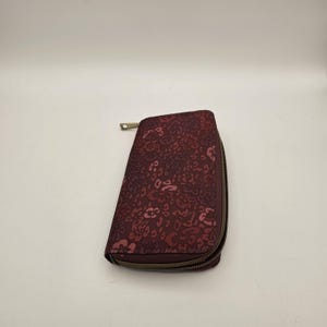 May include: A rectangular zip-around wallet in a deep burgundy color featuring a repeating pattern of stylized floral and leopard-like spots in lighter shades of pink and maroon. The wallet has a metallic zipper closure along the side.