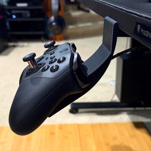 May include: A black gaming controller suspended from a black desk mount. The controller has multiple buttons and two joysticks. The mount is attached to the underside of a black desk. The background includes a wooden floor and other gaming accessories.