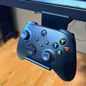 May include: A black Xbox controller with green, yellow, red, and blue buttons is mounted to a black desk mount.