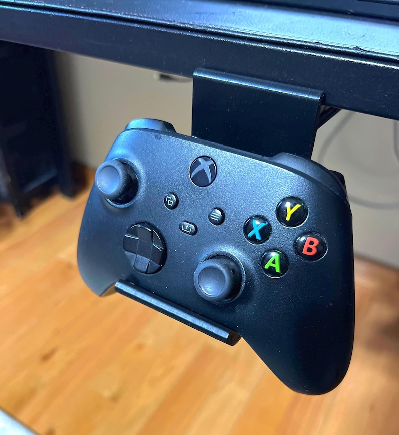 K&ouml;nnte beinhalten: Ein schwarzer Xbox-Controller mit gr&uuml;nen, gelben, roten und blauen Kn&ouml;pfen ist an einer schwarzen Schreibtischhalterung befestigt.