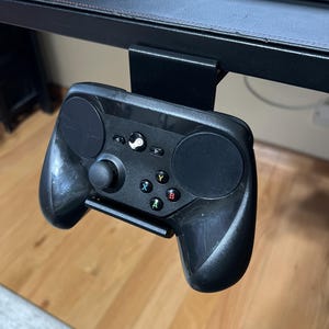 K&ouml;nnte beinhalten: Ein schwarzer Steam-Controller ist mit einer Metallhalterung an einem schwarzen Schreibtisch befestigt. Der Controller hat auf jeder Seite ein kreisf&ouml;rmiges Touchpad, einen Steuerkreuz und Tasten mit den Beschriftungen A, B, X und Y.