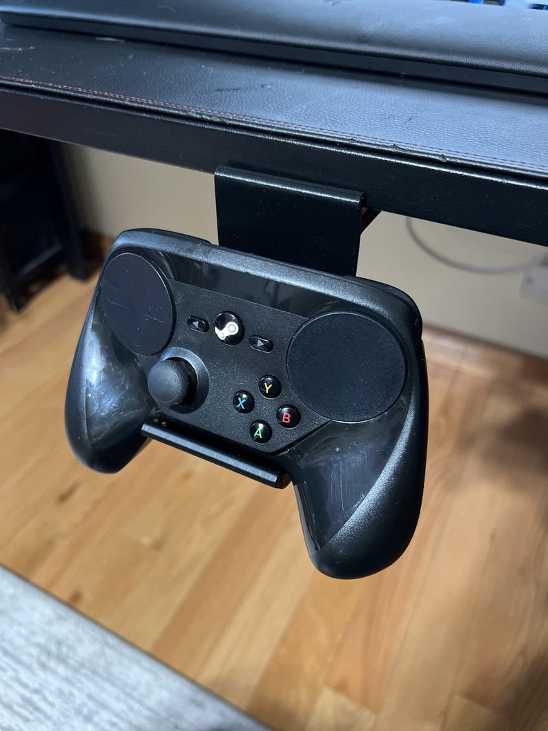 K&ouml;nnte beinhalten: Ein schwarzer Steam-Controller ist mit einer Metallhalterung an einem schwarzen Schreibtisch befestigt. Der Controller hat auf jeder Seite ein kreisf&ouml;rmiges Touchpad, einen Steuerkreuz und Tasten mit den Beschriftungen A, B, X und Y.