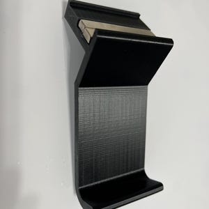 K&ouml;nnte beinhalten: Ein schwarzer 3D-gedruckter Telefonst&auml;nder mit einem integrierten Magnetstreifen. Der St&auml;nder hat eine gebogene Basis und eine erh&ouml;hte Lippe, um ein Telefon an Ort und Stelle zu halten.