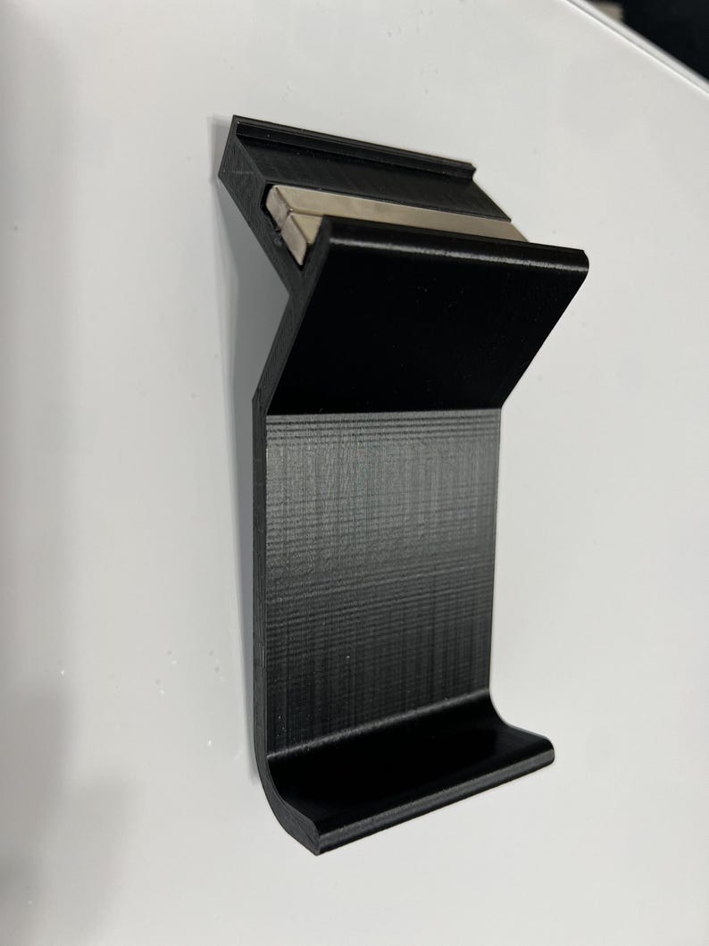 K&ouml;nnte beinhalten: Ein schwarzer 3D-gedruckter Telefonst&auml;nder mit einem integrierten Magnetstreifen. Der St&auml;nder hat eine gebogene Basis und eine erh&ouml;hte Lippe, um ein Telefon an Ort und Stelle zu halten.