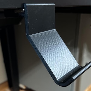 K&ouml;nnte beinhalten: Ein schwarzer, aus Kunststoff gefertigter Haken f&uuml;r die Schreibtischorganisation mit einer strukturierten Oberfl&auml;che. Der Haken ist so konzipiert, dass er unter einen Schreibtisch oder ein Regal geh&auml;ngt werden kann.