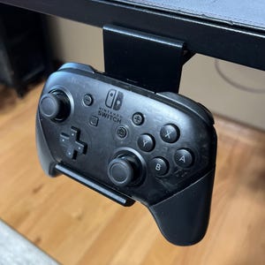 K&ouml;nnte beinhalten: Ein schwarzer Nintendo Switch-Controller ist an einer schwarzen Metall-Tischhalterung montiert. Der Controller hat das Nintendo Switch-Logo und Tasten mit den Beschriftungen A, B, X, Y und +.