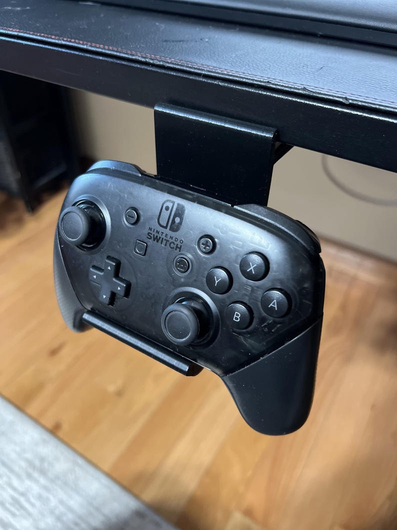 K&ouml;nnte beinhalten: Ein schwarzer Nintendo Switch-Controller ist an einer schwarzen Metall-Tischhalterung montiert. Der Controller hat das Nintendo Switch-Logo und Tasten mit den Beschriftungen A, B, X, Y und +.