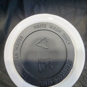 Op de afbeelding: Een witte en zwarte deksel voor een beker. Op de deksel staan de woorden "HAND WASH ONLY", "COFFEE TEA WATER" en "STAINLESS STEEL". De deksel heeft ook een recycling symbool en een logo.