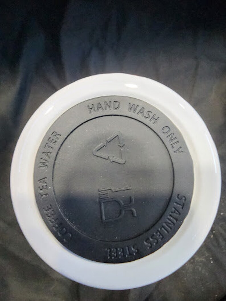 Op de afbeelding: Een witte en zwarte deksel voor een beker. Op de deksel staan de woorden "HAND WASH ONLY", "COFFEE TEA WATER" en "STAINLESS STEEL". De deksel heeft ook een recycling symbool en een logo.