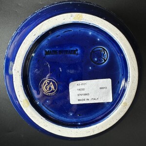 May include: Bottom view of a cobalt blue ceramic bowl with a white rim. The base features the text "MADE IN ITALY" and a sticker with product information. A gold emblem is also visible.