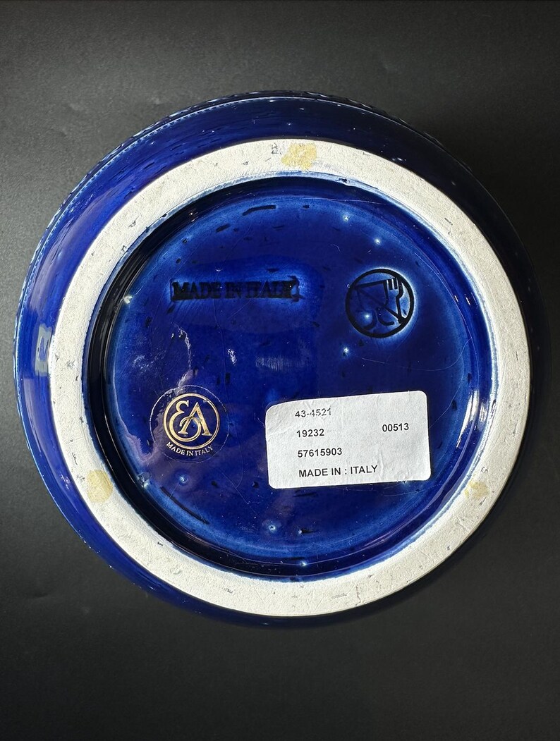 May include: Bottom view of a cobalt blue ceramic bowl with a white rim. The base features the text "MADE IN ITALY" and a sticker with product information. A gold emblem is also visible.