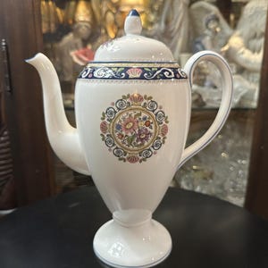 May include: White ceramic coffee pot with a lid and handle. The pot features a floral design in the center and a decorative border around the top. The spout and handle are curved. The pot is on a pedestal base.