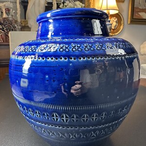 May include: A large, cobalt blue ceramic vase with a lid. The vase features a textured surface with geometric patterns and perforations. The glossy finish reflects light, highlighting the intricate design and shape. A decorative piece for home decor.