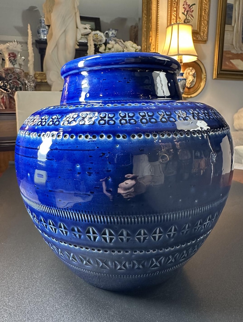 May include: A large, cobalt blue ceramic vase with a lid. The vase features a textured surface with geometric patterns and perforations. The glossy finish reflects light, highlighting the intricate design and shape. A decorative piece for home decor.