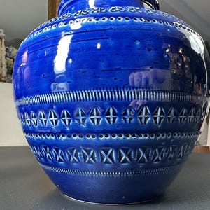 May include: A large, cobalt blue ceramic vase with intricate geometric patterns. The vase features a wide, rounded body with a narrow neck and a series of decorative bands. The surface has a glossy finish, reflecting light and highlighting the detailed design.