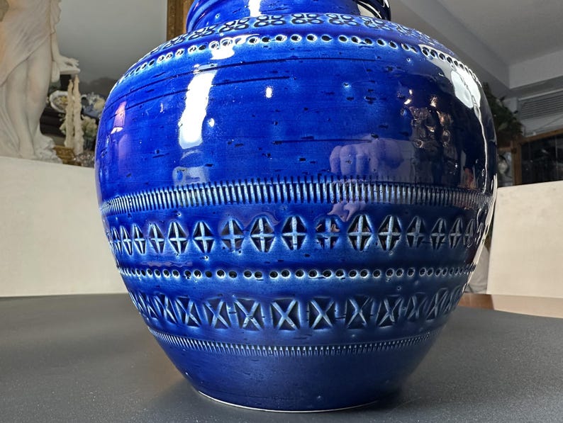 May include: A large, cobalt blue ceramic vase with intricate geometric patterns. The vase features a wide, rounded body with a narrow neck and a series of decorative bands. The surface has a glossy finish, reflecting light and highlighting the detailed design.