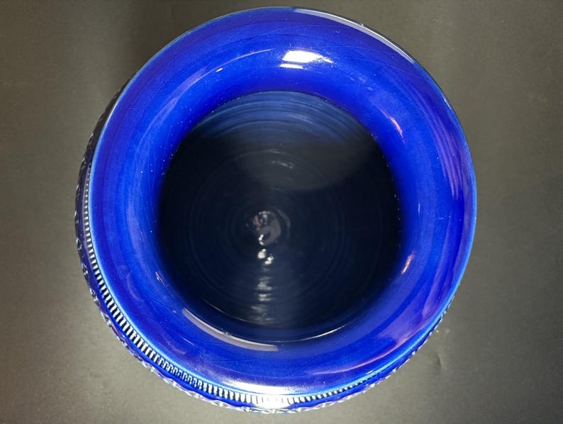May include: A top-down view of a deep blue ceramic vase. The vase has a wide, rounded rim and a textured interior. The rim is a brighter shade of blue than the interior. The vase has a decorative pattern around the outside.