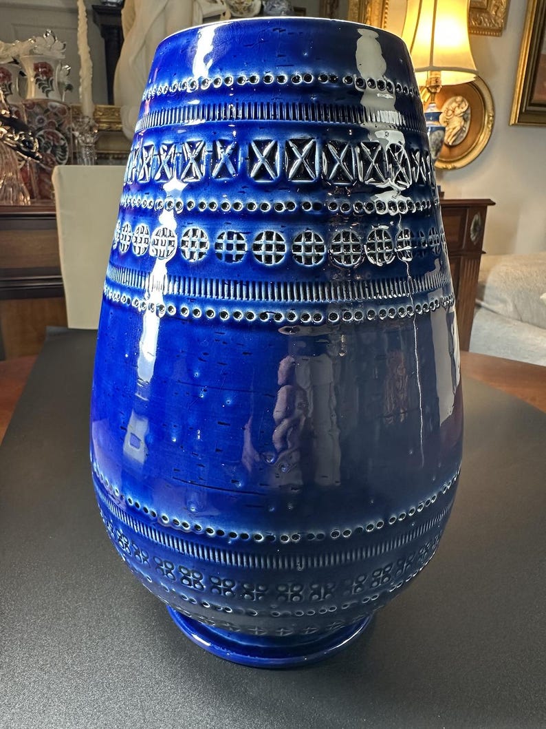 May include: A large, cobalt blue ceramic vase with a glossy finish. The vase features intricate, geometric patterns and perforations around the body. The design includes rows of circles, crosses, and other shapes. The vase has a wide base and a slightly flared top.