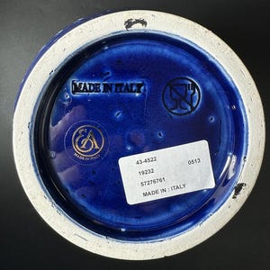 May include: Close-up of the base of a ceramic vase. The interior is a deep blue with a white rim. The base has the text "MADE IN ITALY" and a sticker with numbers. The vase has a glossy finish.