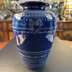 May include: A tall, cobalt blue ceramic vase with intricate white embossed patterns. The vase features a wide base, a cylindrical body, and a slightly flared rim. The patterns include geometric shapes and dotted lines, creating a textured surface.