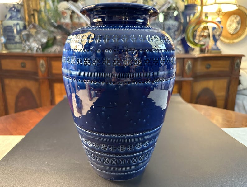 May include: A tall, cobalt blue ceramic vase with intricate white embossed patterns. The vase features a wide base, a cylindrical body, and a slightly flared rim. The patterns include geometric shapes and dotted lines, creating a textured surface.