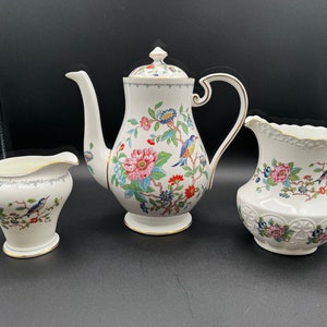 Puede incluir: Un juego de té de porcelana blanca que incluye una tetera, una jarra para la crema y un azucarero. El juego está decorado con motivos florales y de aves coloridas, con detalles dorados. La tetera tiene un pico largo y curvo y una tapa con un pomo.