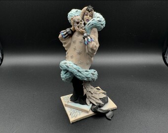 May include: A porcelain figurine of a woman in a white dress with a blue fur collar, holding a skull in her hands. The figurine is standing on a white base with a floral design.