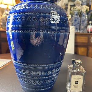 May include: A large, cobalt blue ceramic vase with a glossy finish. The vase features intricate embossed patterns and a wide, rounded body. A clear Jo Malone cologne bottle is next to the vase.