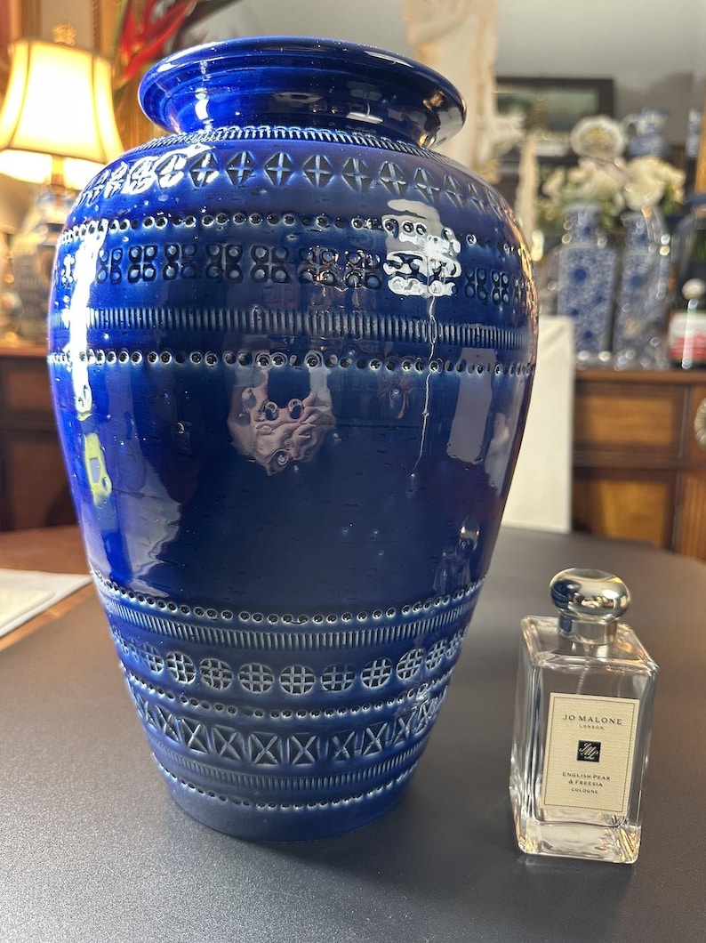 May include: A large, cobalt blue ceramic vase with a glossy finish. The vase features intricate embossed patterns and a wide, rounded body. A clear Jo Malone cologne bottle is next to the vase.